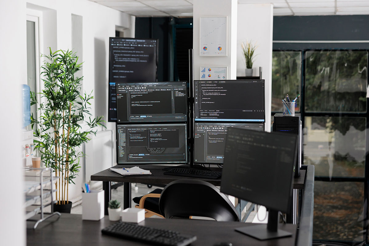 An empty open-plan IT office with a bank of four monitors on a stand showing code editors, a second monitor in the foreground, a bamboo plant and a desk with paperwork