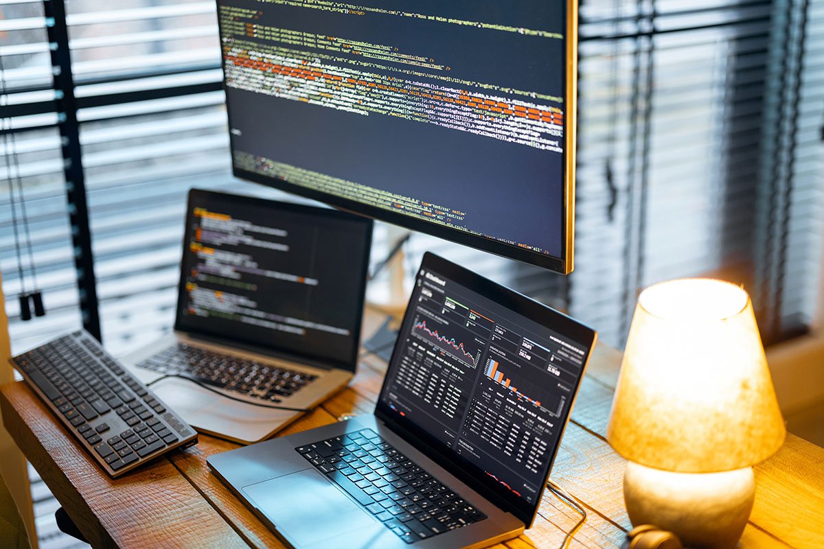A wooden desk with a large monitor and laptops showing source code and a dark analytics dashboard beside a lit table lamp