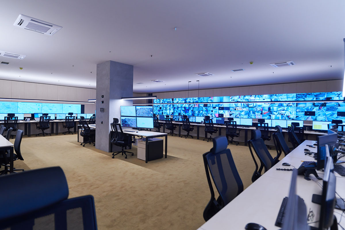An empty modern security operations centre with rows of desks and a wall of monitors displaying surveillance and network feeds