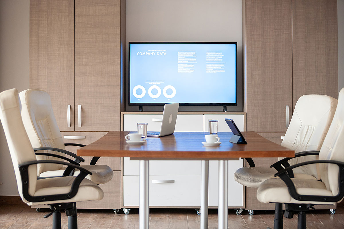 An empty meeting room with white leather chairs around a wooden table and a wall screen showing a company data presentation