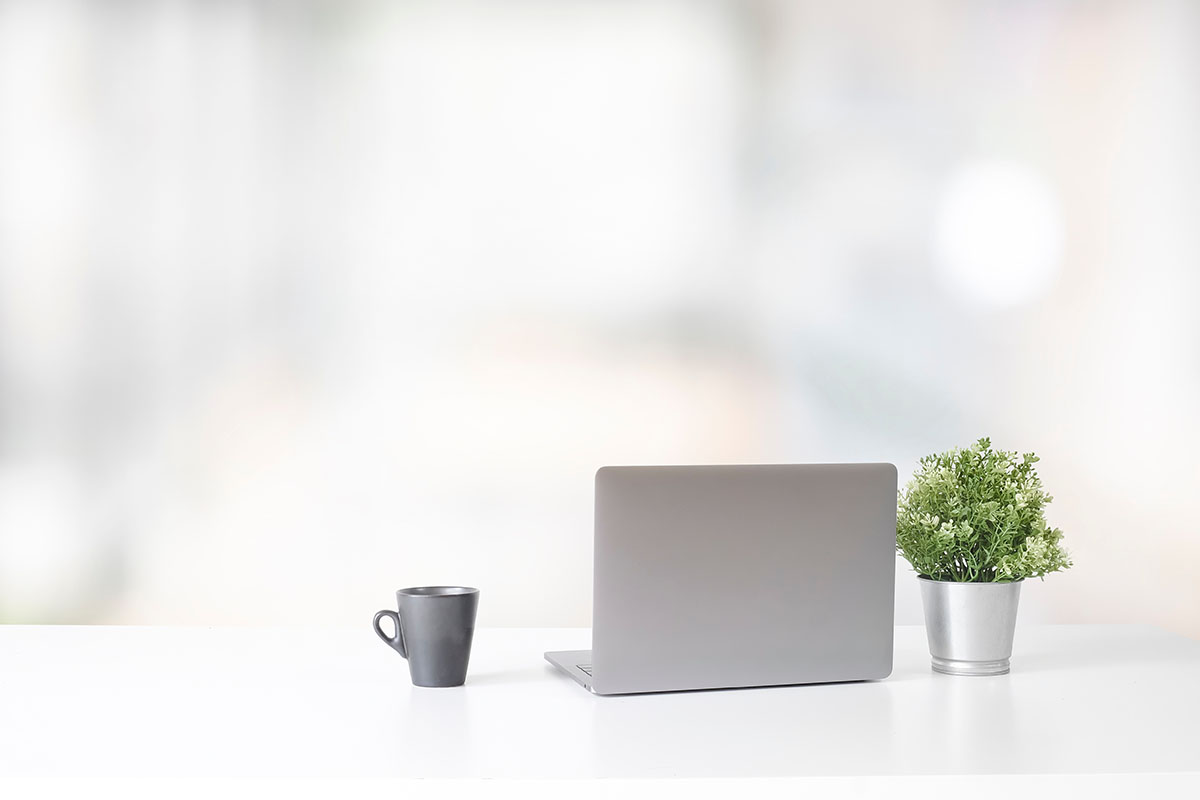 A closed laptop, coffee cup and small plant on a minimalist white office desk