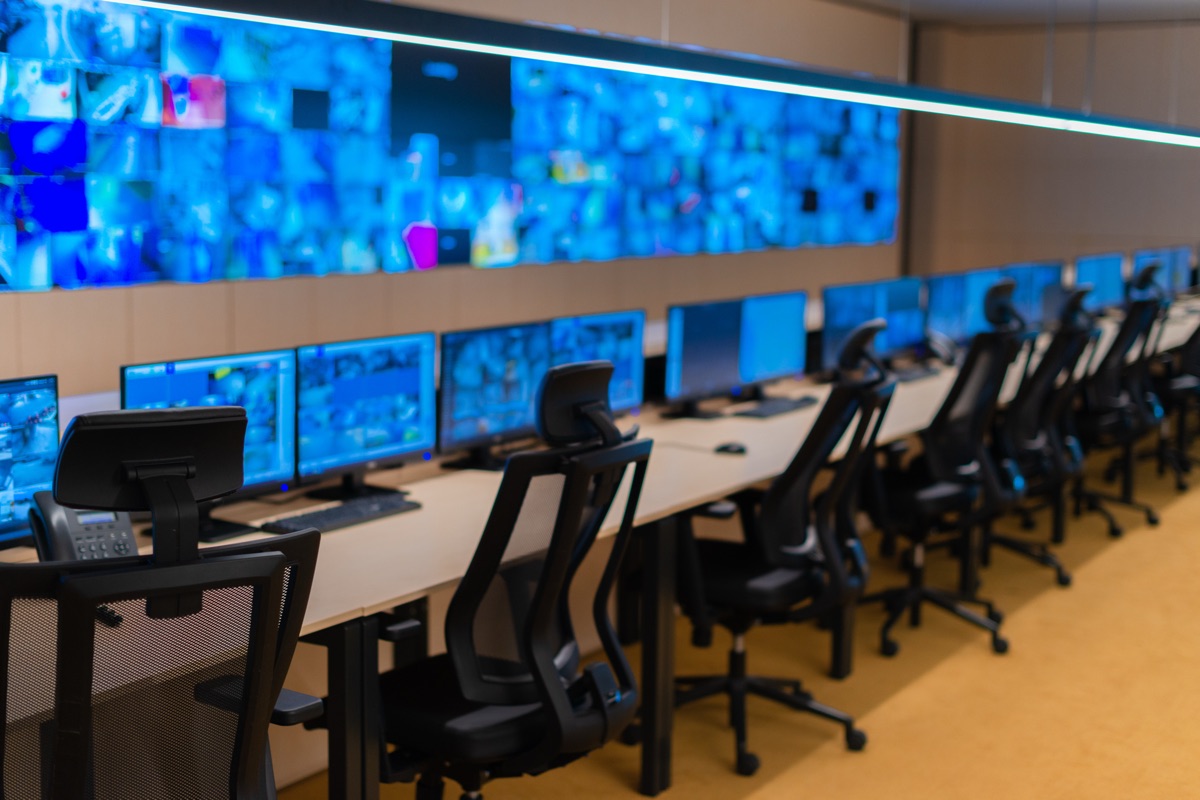 Rows of empty black office chairs facing monitors in a dimly lit operations center, with a wall of blue screens behind them