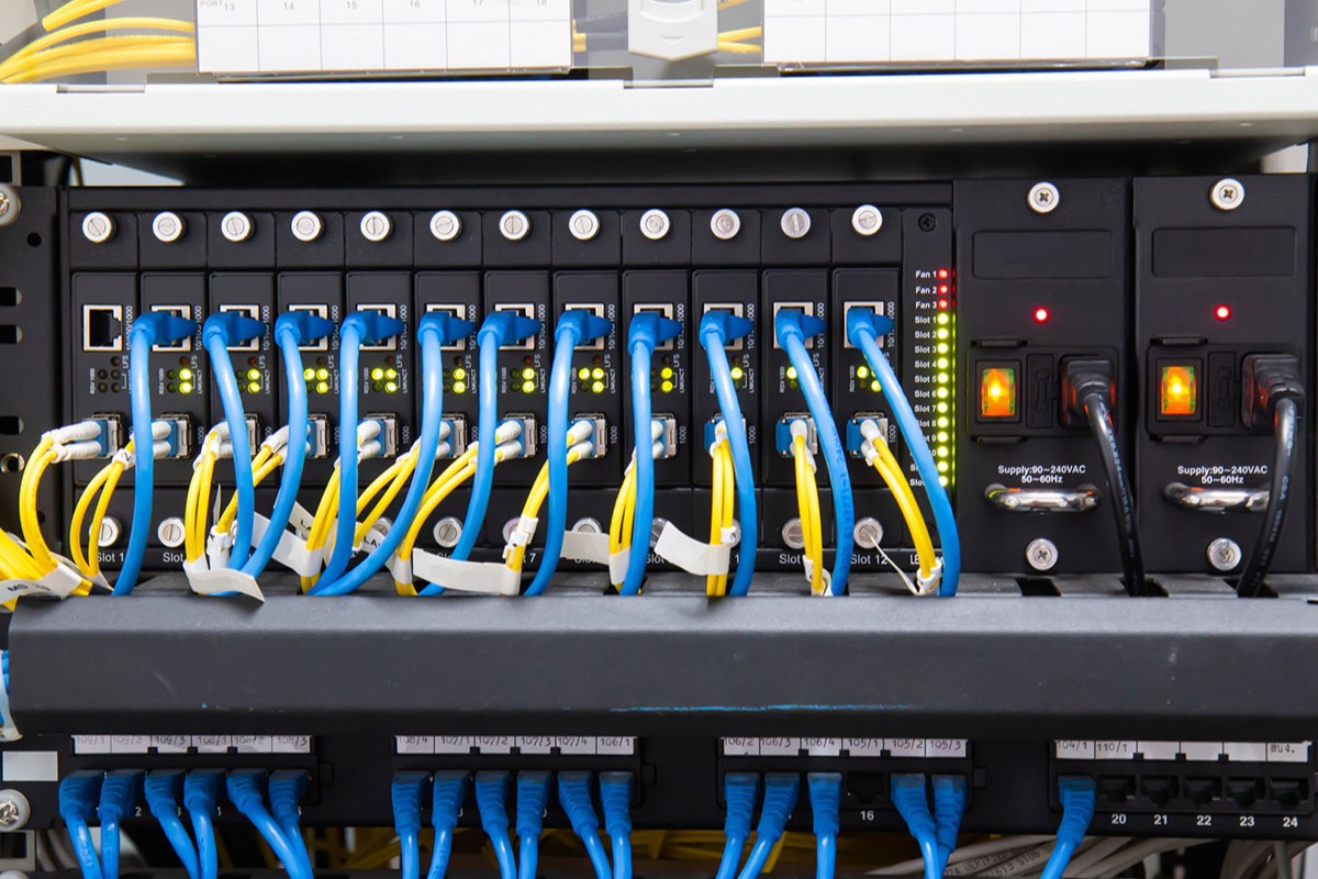 Front of a rack-mounted network chassis with a row of modules connected by blue ethernet cables and yellow fiber patch cords, green status lights and two power supplies with amber switches on the right, and a patch panel of blue cables below