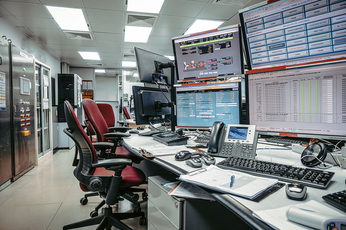 An empty monitoring room with banks of status screens above a curved desk of keyboards, desk phones and paperwork